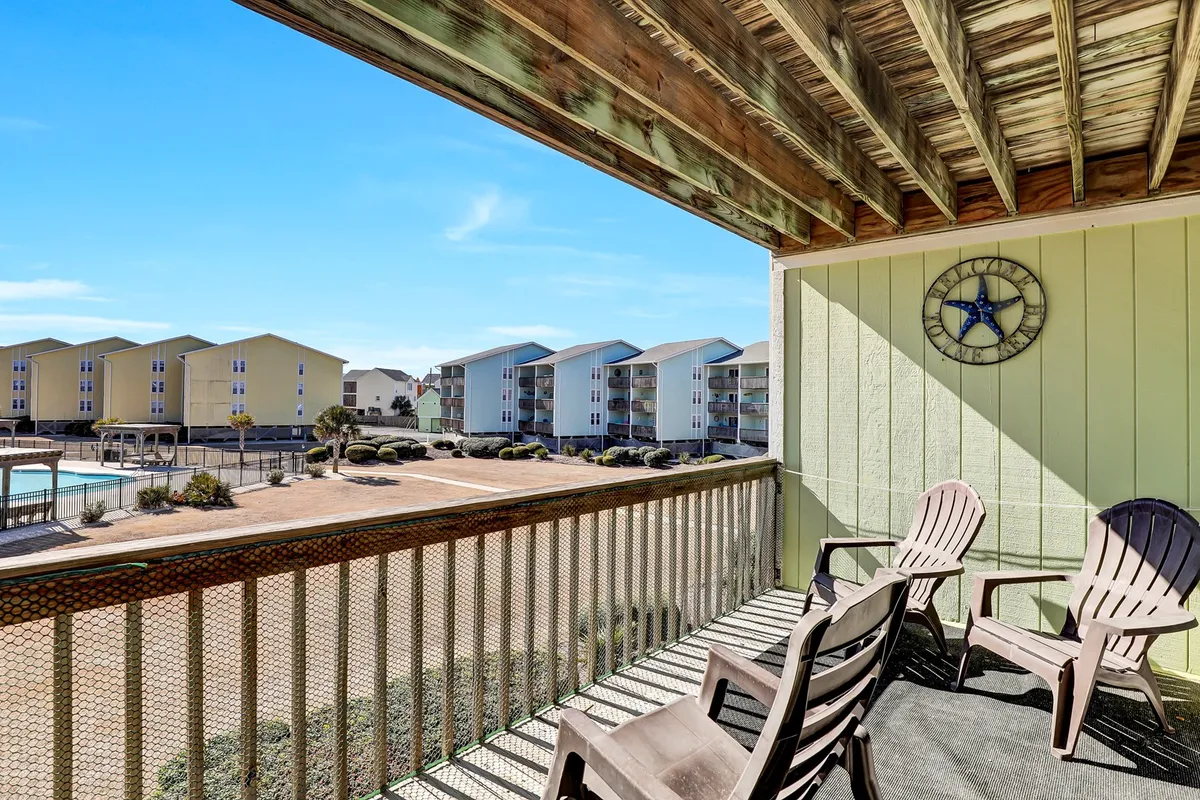 A welcoming balcony scene featuring chairs overlooking the pool area, with surrounding buildings and a prominent clock on the wall. The vantage point provides a sense of the property's layout and hints at the pleasure of watching sunrises or relaxing outdoors while staying close to the beach.