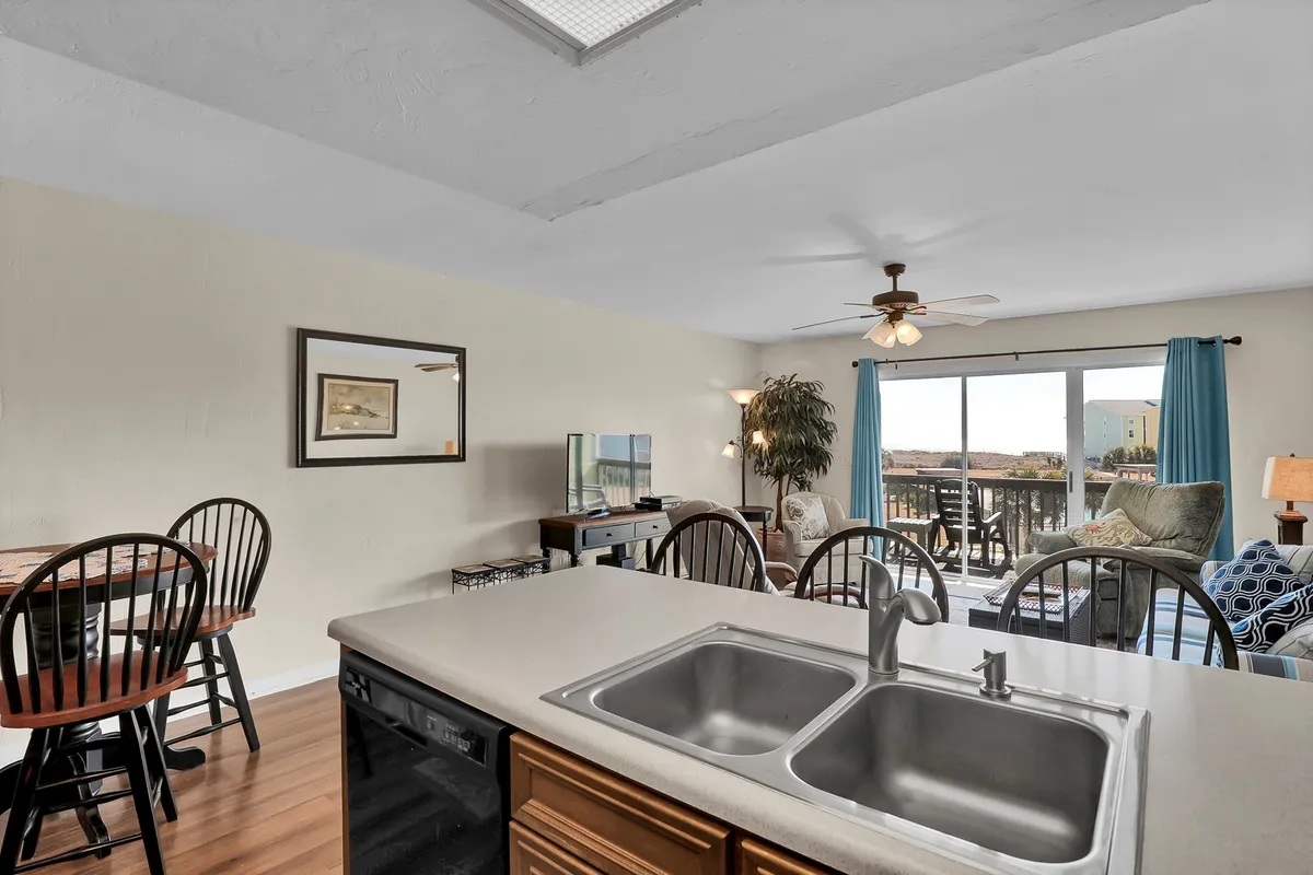 A wide, inviting view of the open-concept living area blends a cozy couch, a dining table, and a kitchen counter, all illuminated by natural light streaming through large windows with scenic views beyond. The space feels airy and designed for both relaxation and togetherness.