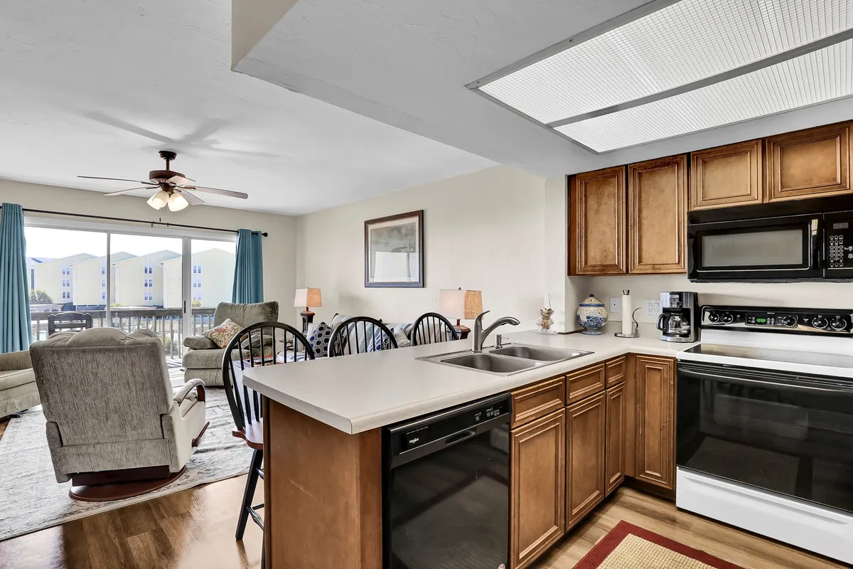 This image presents a welcoming, light-filled interior, highlighting a kitchen counter with sink, black appliances, wood cabinetry, a dining area with chairs, and an adjacent living area bathed in natural light. The space feels open and inviting, with a layout conducive to both relaxing and entertaining.
