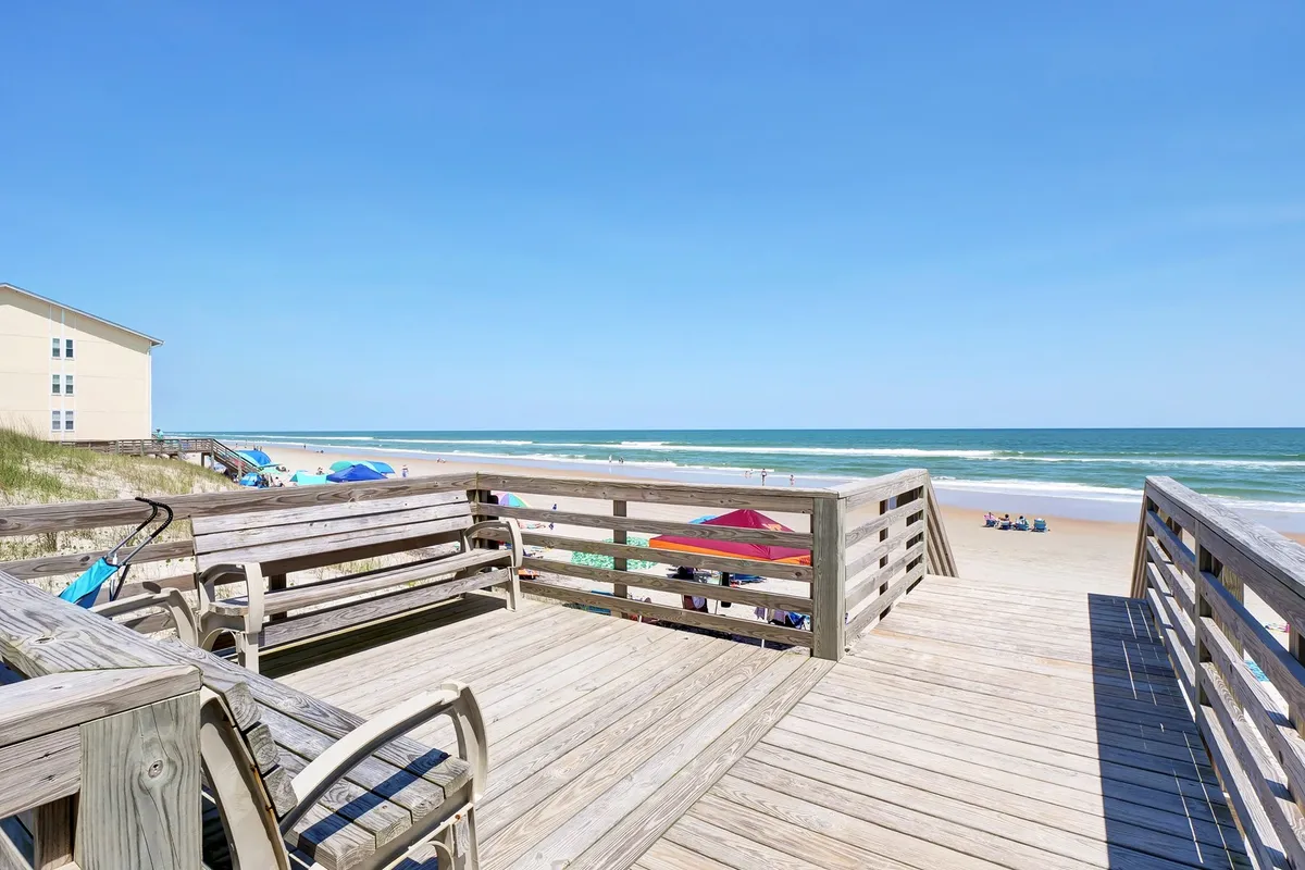 A private deck overlooks the sandy beach and rolling ocean waves, furnished with seating for soaking in the sunrise or sunset. Beachgoers, umbrellas, and a vibrant coastal scene complete the view, connecting the indoors with the outdoors.