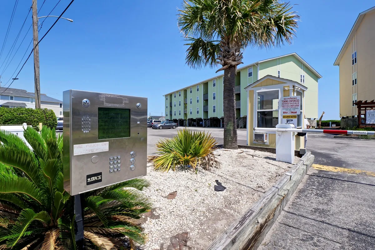 The image presents the exterior entrance to the property, featuring an intercom system, palm trees, an entrance booth, and the buildings beyond. The landscaping and tropical ambiance suggest a welcoming, vacation-oriented environment that encourages exploration beyond the condo itself.