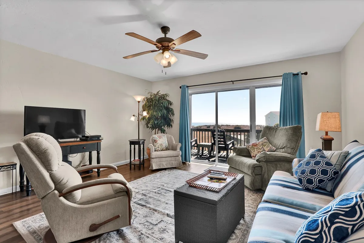 A comfortable living room with a sofa, armchair, television, and a coffee table sits adjacent to large windows offering a view outside. The arrangement invites relaxation, socializing, and catching glimpses of the ocean or sunsets from indoors.