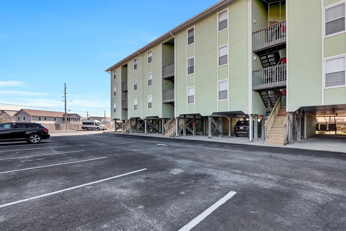 This exterior shot highlights the property's green building, parking area, and staircase, subtly indicating easy access to the beach from the building itself. The casual mood and community vibes underscore the convenience of coming and going for beach adventures.