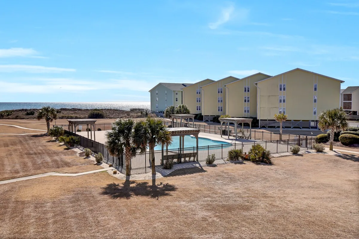 A vibrant outdoor pool scene framed by swaying palm trees and yellow resort-style buildings, with a hint of the ocean visible in the background. Sun loungers and poolside areas invite guests to swim, sunbathe, and gather for outdoor fun.