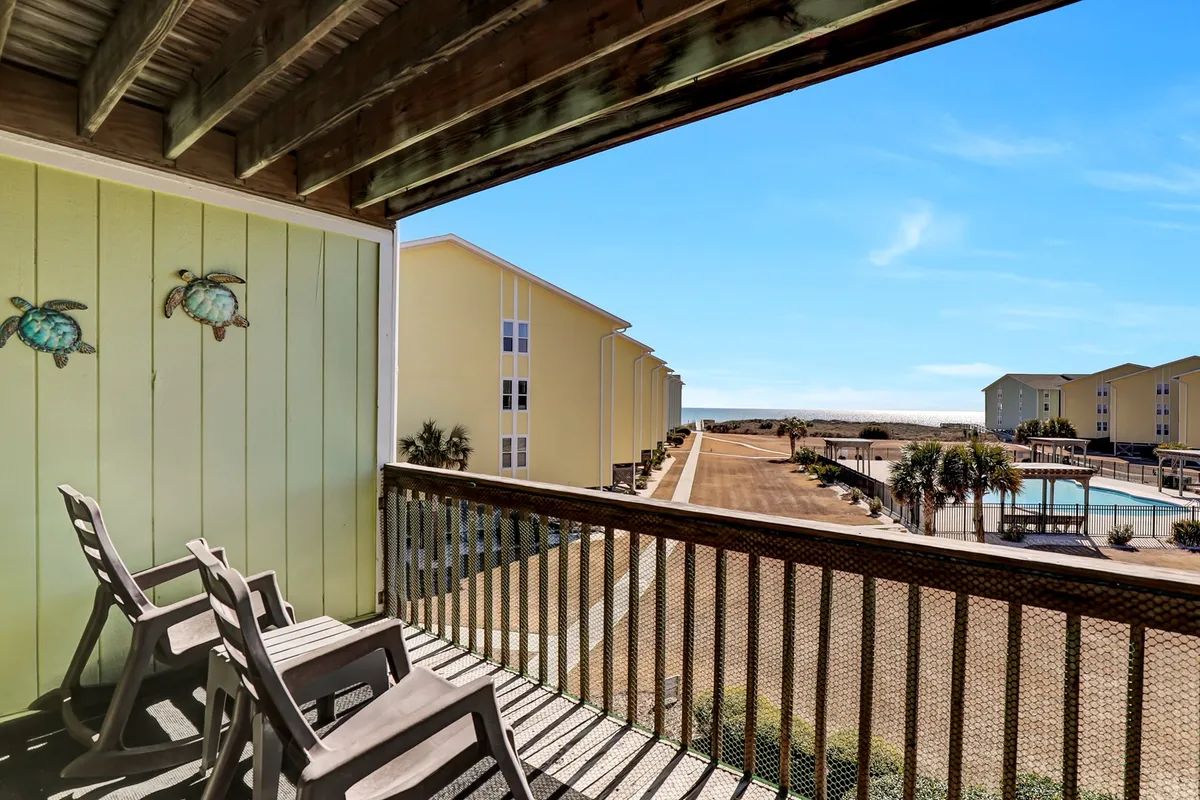 The image showcases the exterior of a charming Surf City condo, featuring a private balcony with inviting chairs adorned by whimsical turtle decorations. Below, a sparkling pool area beckons guests to relax or gather with friends, all set against a tranquil coastal backdrop ideal for sunrise views or evening unwinding.