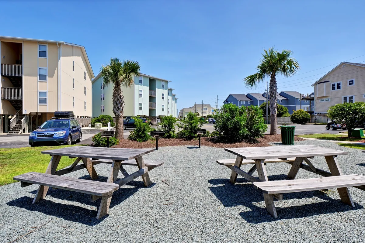 A tranquil outdoor setting featuring wooden picnic tables shaded by palm trees. The landscaped bushes and open grassy areas create an inviting space for gatherings, meals, and relaxation under the sun, suggesting opportunities for both family fun and quiet relaxation.